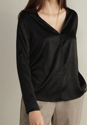 Femme portant un chemisier noir à manches longues et col en V et un pantalon beige, debout devant un fond uni clair.