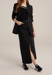 Svart denimkjol med slits fram, tillsammans med en svart blazer. Svarta sneakers har vita ränder och en blå logodetalj.