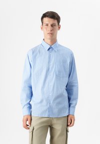 Un joven está de pie contra un fondo simple, vistiendo una camisa de rayas azul claro y pantalones beiges, con una expresión neutral.