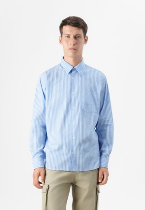 A young man stands against a plain background, wearing a light blue striped shirt and beige pants, with a neutral expression.