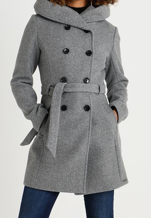 Kvinde iført en grå dobbeltradet uldfrakke med bælte, hætte og hænder i lommerne, kombineret med mørkeblå jeans.