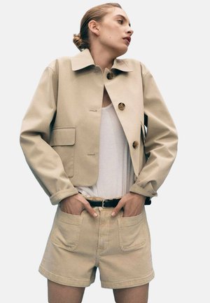 Beige cropped jakke med store knapper, frontlommer og en flad krave, parret med lyse beige shorts og en hvid T-shirt. Glat tekstur.