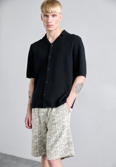 Schwarzes Kurzarm-Hemd mit Knöpfen aus weichem Strickstoff, kombiniert mit hellen Shorts mit geometrischem Muster.