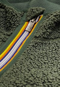 Giacca in pile di colore verde scuro con una superficie texturizzata, caratterizzata da una zip multicolore con strisce gialle, arancioni, viola e bianche.