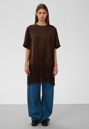 Femme debout face à l'avant, porte un haut tunique ample marron à franges, un large jean bleu et des chaussures noires, sur fond clair et uni.