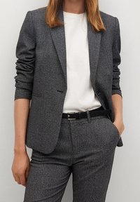 Blazer à carreaux gris avec manches longues, fermeture à un seul bouton et texture subtile, porté sur une chemise blanche et une ceinture noire. Le pantalon assorti complète la tenue.