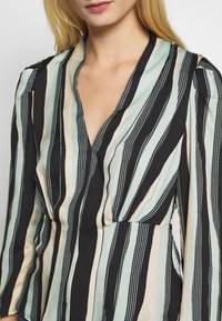 Czarny i kremowy top w pionowe paski o różnych szerokościach, zdobiony jasnoniebieskimi i białymi kropkami. Dekolt w serek i długie rękawy.