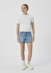 Camiseta blanca de manga corta combinada con pantalones cortos de mezclilla azul. El modelo lleva zapatillas blancas y calcetines con estampado. Diseño simple y casual.