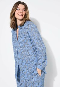 Blauwe shirt met paisleypatroon, met kraag, knoopsluiting aan de voorkant, lange mouwen en zijzakken; de stof lijkt zacht en lichtgewicht te zijn.
