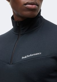 Κοντινό πλάνο ενός ατόμου που φοράει μαύρο τοπ με φερμουάρ και ψηλό γιακά, με λευκό λογότυπο "Peak Performance" στο στήθος.