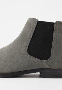 Bota de tornozelo em camurça cinza com painéis laterais elásticos e um inserto preto. Sola plana com acabamento texturizado, apresentando detalhes de costura mínimos.