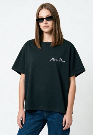 Femme aux cheveux bruns et raides, portant des lunettes de soleil noires en forme d'œil de chat, un t-shirt noir ample avec l'inscription "Mira Paris" et un jean bleu sur un fond uni.