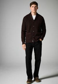 Brun, texturerad cardigan med sjalkrage, sex knappar och två framfickor, kombinerad med svarta byxor och bruna skor.
