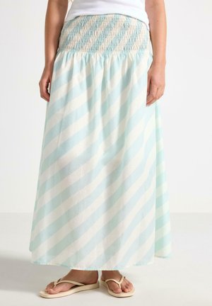 Kvinde iført en lang, lyseblå og hvid diagonalt striped nederdel med smocktalje og beige flip-flops på en hvid baggrund.