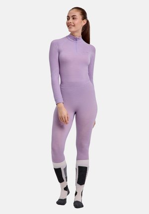 Jonge vrouw in een lila aansluitend langemouwshirt en legging met rits bij de hals, draagt witte en zwarte kniekousen, staand en glimlachend.