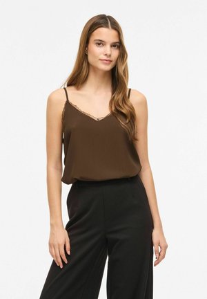 Jeune femme aux longs cheveux portant un haut caraco marron avec finitions en dentelle et un pantalon noir taille haute, debout sur un fond blanc.