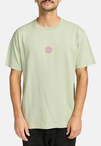 Lichtgroene katoenen t-shirt met een centraal grafisch ontwerp van een roze zon met de letters "RVCA" eromheen. Korte mouwen, losse pasvorm.