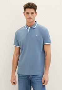 Blå polo t-shirt i bomull, med krage med kontrasterande vita ränder och en liten logotyp på bröstet. Kort ärm, enkel design.