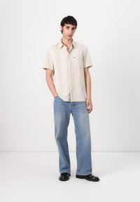 Kortermet lys beige skjorte med knapper og vertikale striper, sammen med løstsittende blå jeans og svarte sko.