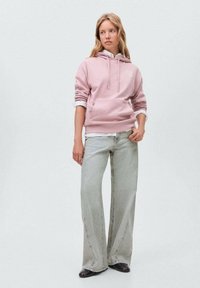 Rosa hoodie med en framficka, snören för huva och vit text. Kombinerad med ljusgrå utsvängda jeans och svarta skor.