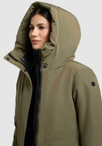 Olivgrüne isolierte Jacke mit einer großen Kapuze, Reißverschluss, schwarzen Akzenten und Druckknöpfen. Verfügt über einen strukturierten, wasserabweisenden Stoff.