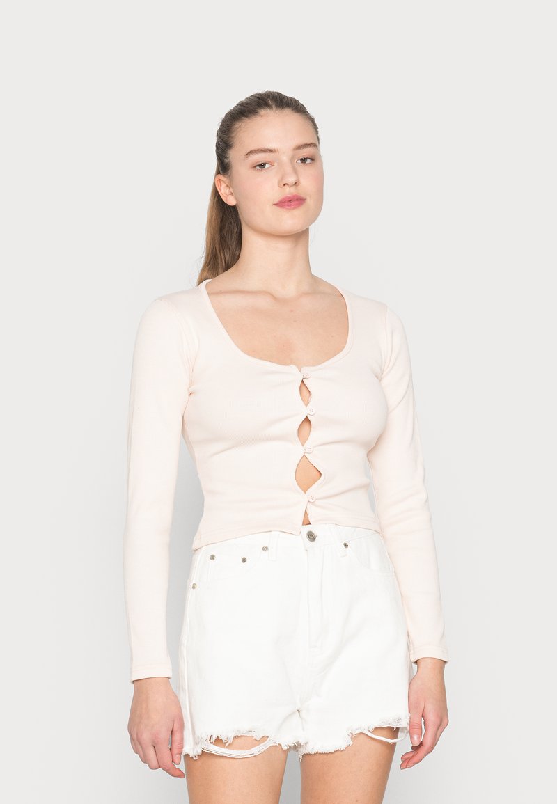 Top de manga longa rosa claro com um decote em U profundo, apresentando cinco botões decorativos em forma de diamante ao centro, combinado com calções brancos.