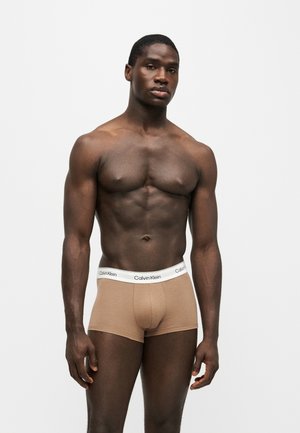 Muskelöser Mann trägt beige Calvin Klein Boxershorts mit weißem Bund, steht vor hellem einfarbigem Hintergrund.