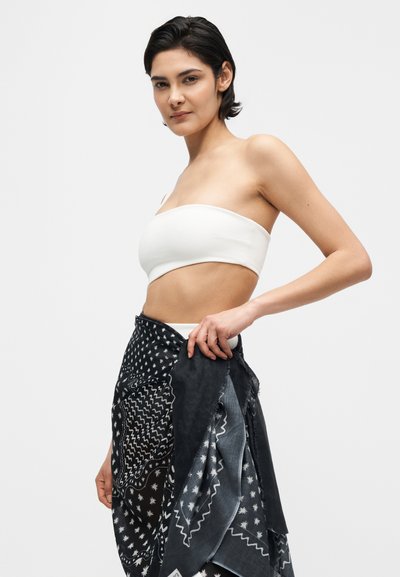 Γυναίκα με κοντά σκουρόχρωμα μαλλιά, που φοράει λευκό strapless τοπ και τυλίγει μια μαύρη φαρδιά μαντήλα με σχέδια γύρω από τη μέση της, σε λευκό φόντο.