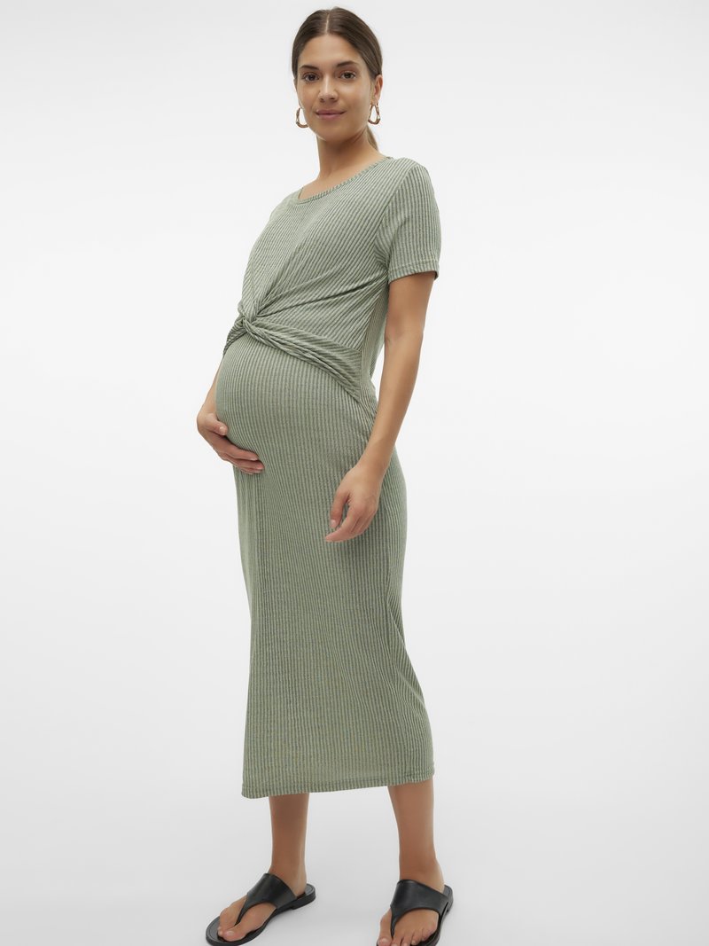 Pregnant woman wearing a ribbed, short-sleeve, sage green midi dress with a front knot detail, holding her belly and standing in black sandals.