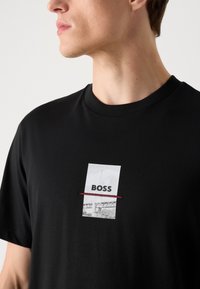 Melna kokvilnas T-kreklā ar centrālu grafiku baltā krāsā, kurā ir izdrukāts "BOSS", akcentēts ar sarkanu līniju. Īsām piedurknēm, apaļš kakls.