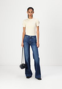 Kremfarget ribbete topp med korte ermer, kombinert med høyt liv mørkeblå vide jeans. Stående posisjon, holder en svart rund veske.