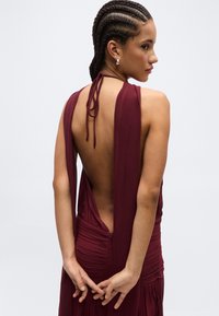 Kvinde med fletninger iført en ærmeløs, rygfri vinrød kjole, vendt sidelæns med hænderne foldet bag ryggen.