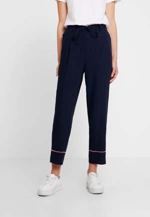 Frau trägt marineblaue, gekürzte Hose mit Taillenband und weißen Turnschuhen, steht vor einem schlichten hellen Hintergrund.