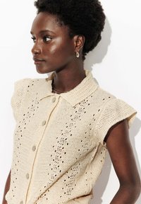 Haut en tricot beige clair à boutons avec des manches courtes à volants et des détails en œillets. Il présente un col classique et des boutons ronds.