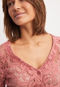 Pink crochet cardigan featuring a v-neck design, button closure, and intricate lace patterns, showcasing a lightweight and textured knit style.