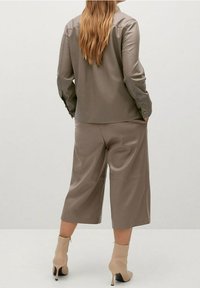 Achteraanzicht van een model dat een lichtbruine blouse met knopen draagt en bijpassende wijde broekspijpen, gecombineerd met beige enkellaarsjes met hoge hakken.