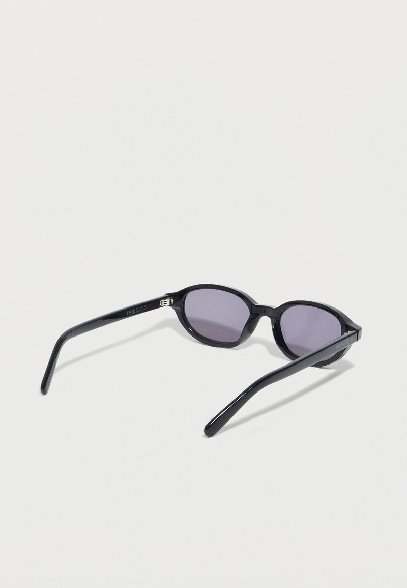 Gafas de sol ovaladas negras con lentes de tono oscuro, abiertas y apoyadas sobre una superficie blanca, orientadas para mostrar el interior y los detalles del lado izquierdo.