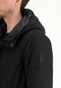 Giacca nera con cappuccio, dotata di un rivestimento interno trapuntato, con chiusura zip frontale e un'etichetta logo nera testurizzata sulla manica sinistra.