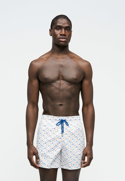 Μυώδης άνδρας με ουδέτερη έκφραση, φορώντας λευκά μαγιό σορτς με πολύχρωμα σχέδια αστεριών και μπλε κορδόνι, μπροστά σε απλό φόντο.