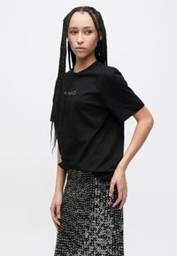 Črna majica s kratkimi rokavi z napisom "PINKO", kombinirana s črno krilo, okrašeno s srebrnimi okroglimi dodatki, ki ustvarjajo teksturiran vzorec.