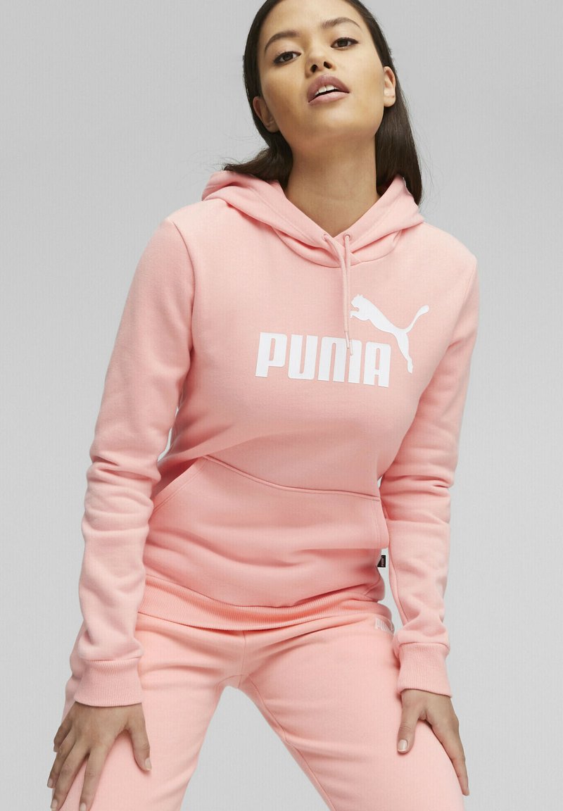 Felpa rosa con logo "PUMA" bianco, caratterizzata da una tasca a marsupio e polsini a costine. Abbinata a pantaloni della tuta rosa coordinati, che mostrano un design rilassato.