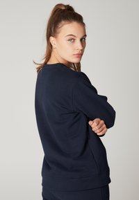 Felpa di un blu navy scuro in tessuto morbido con maniche lunghe, collo a giro e polsini a costine. La vista laterale mette in evidenza la vestibilità rilassata e il design semplice.