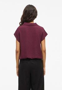 Short-sleeve, burgundy top with a textured finish and a collar, paired with black pants. The top has a relaxed fit and falls at the waist.
