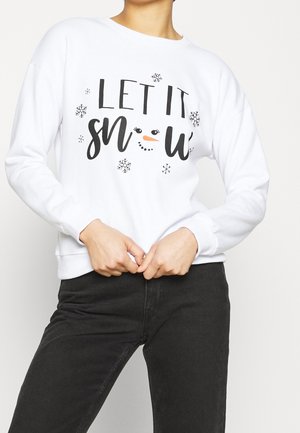 Personne portant un sweat-shirt blanc avec le texte noir "LET IT SNOW" et des motifs de flocons de neige, assorti à un pantalon noir.
