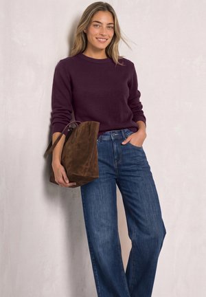 Femme souriante portant un pull violet et un jean bleu à jambes larges, tenant un grand sac fourre-tout en daim marron, debout contre un mur clair.