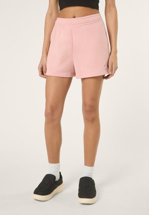 Vrouw die lichtroze shorts met elastische taille draagt, zwarte instapschoenen en witte sokken, staand tegen een effen lichte achtergrond.