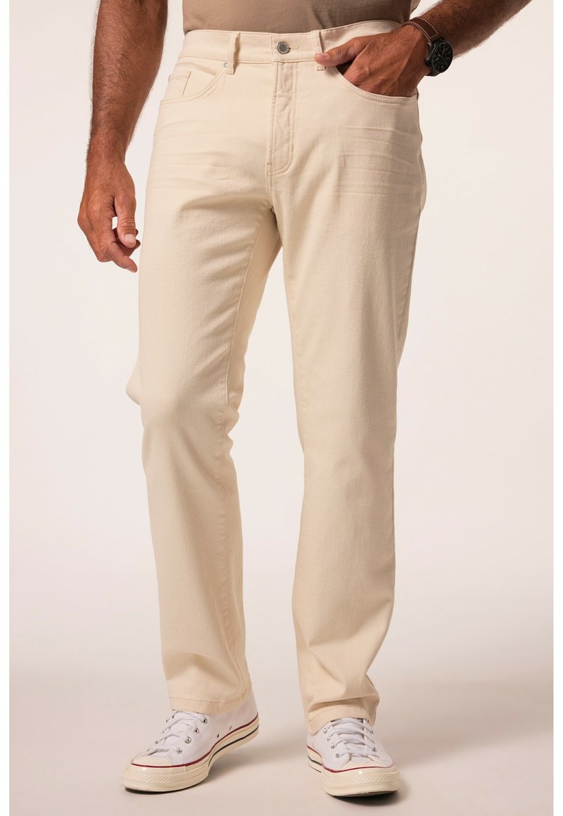 Beige Baumwoll-Chinos mit geradem Bein, vorderen Taschen und einem Klemmverschluss. Kombiniert mit weißen Sneakers und einem taillierten hellbraunen Hemd.