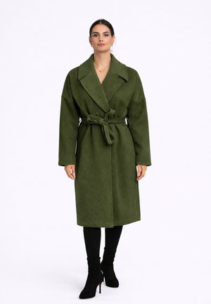 Kobieta stoi ubrana w oliwkowo zielony wełniany płaszcz z paskiem i szerokimi klapami, w połączeniu z czarnymi legginsami i czarnymi botkami na obcasie.