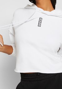 Bílá crop mikina s kapucí na stažení a vertikálním černým logem "HUGO" na přední straně, doplněná černými kalhotami.