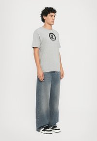 Tricou gri din bumbac cu mâneci scurte și un design grafic circular negru pe piept, asociat cu blugi evazați decolorati de culoare albastră și adidași.